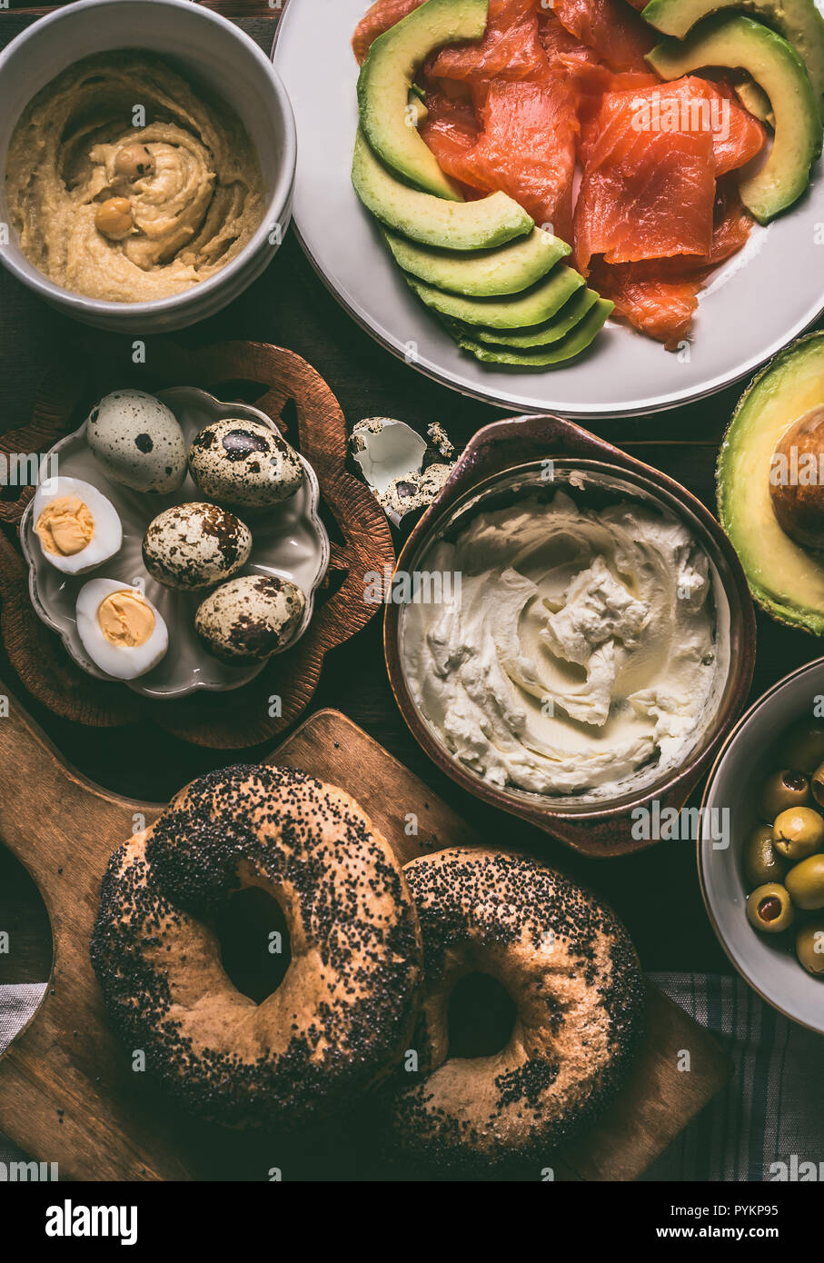 Breakfast preparation with bagel bread, salmon, avocado , fresh cheese, hummus and cooked quail eggs on dark rustic wooden background, top view. Stock Photo