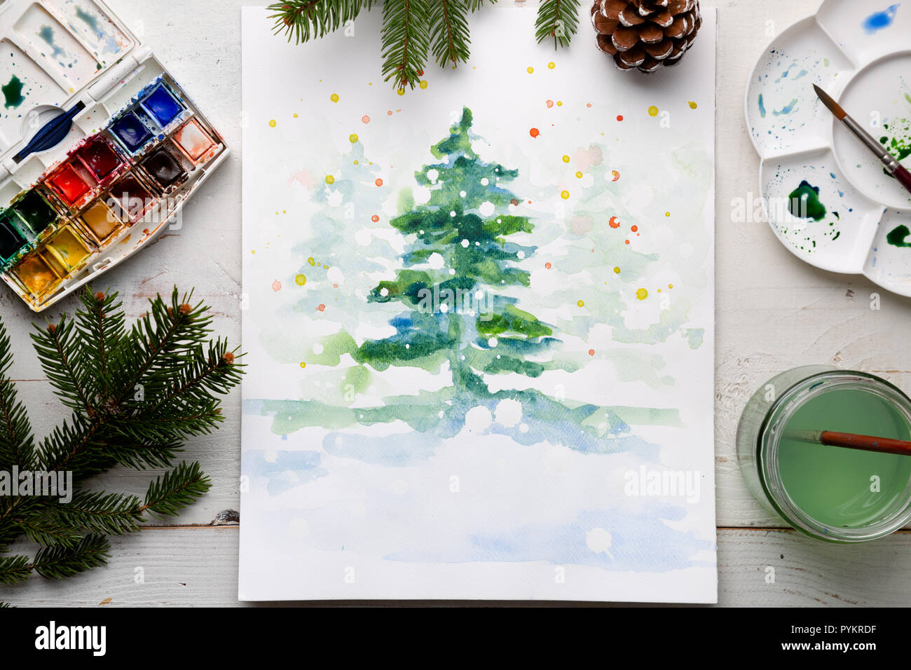 Handmade watercolor Christmas card on the work table with watercolor box, brushes, palette and fir branches and pine cones. Top view Stock Photo