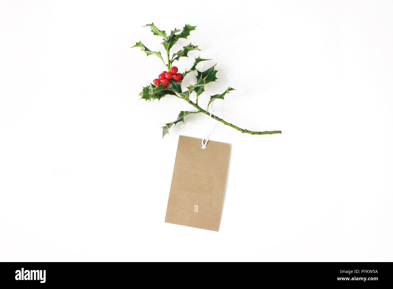 Close-up of craft paper gift tag with rope and green holly branch with red berries isolated on white table background. Christmas composition, top view. Stock Photo
