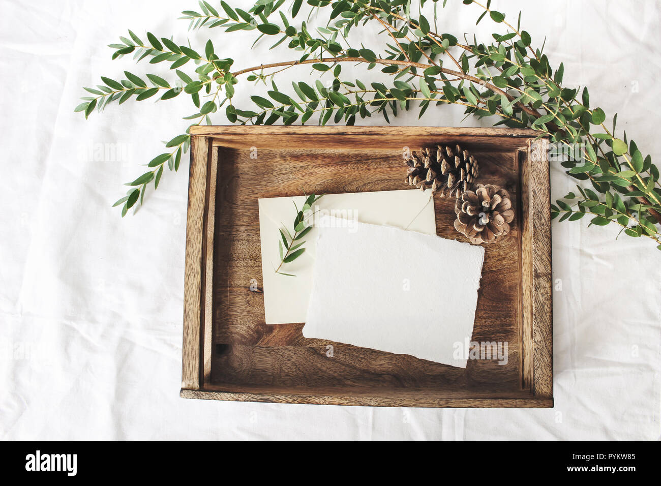 Christmas or winter wedding mock-up scene. Blank cotton paper greeting cards, old wooden tray, pine cones and green Eucalyptus parvifolia branch.White bed linen background. Flat lay, top view. Stock Photo