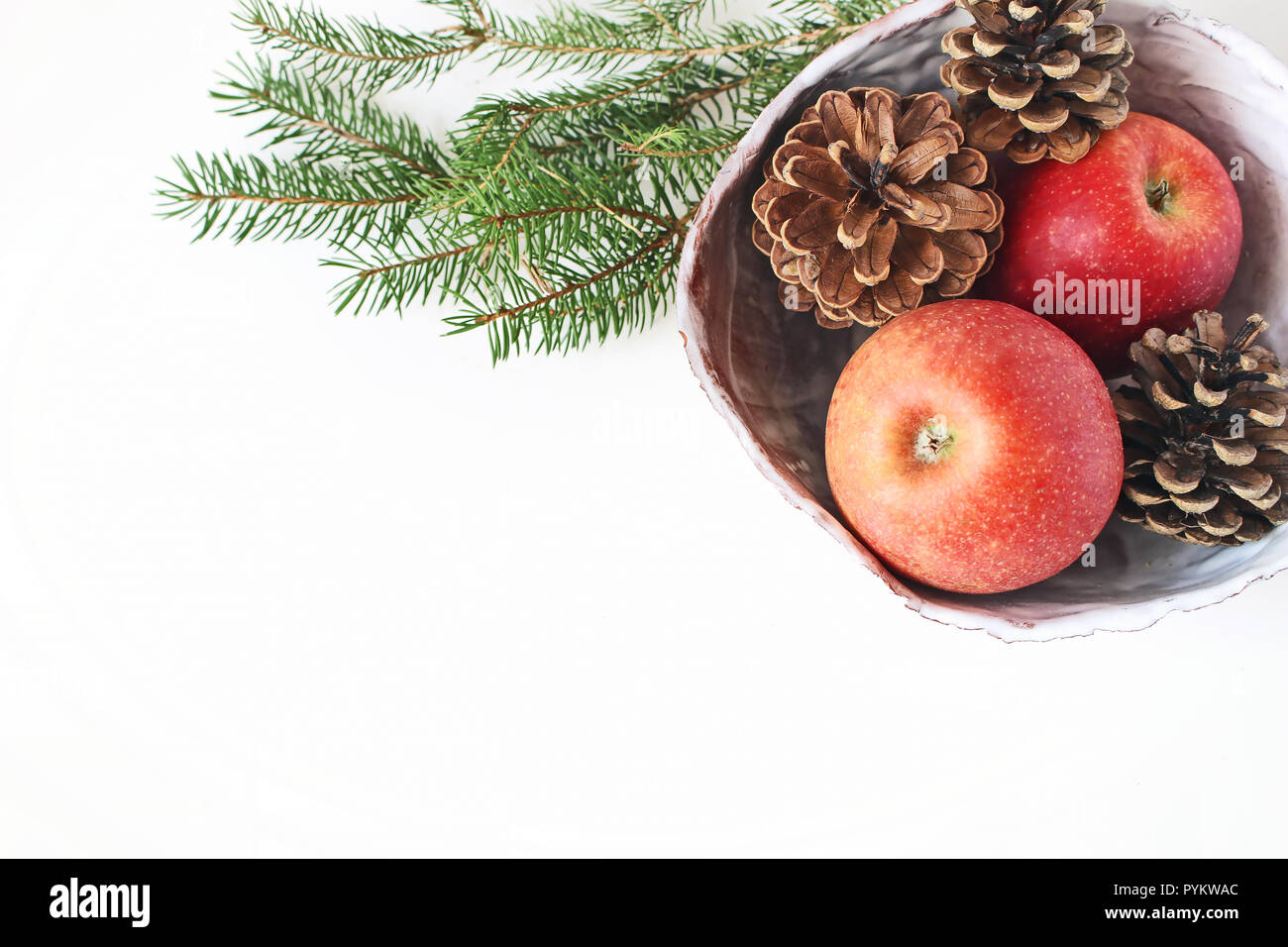 Closeup of red apple fruit and pine cones in ceramic bowl and spruce tree branches on white table background. Christmas holiday styled stock photo. Winter food composition. Flat lay, top view. Stock Photo