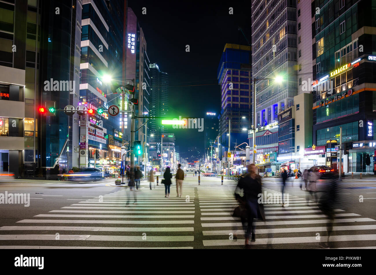 People cross a pedestrian crossing at night in Haeundae, Busan, South Korea. Behind, the street is full or bright lights shining in the dark. Stock Photo