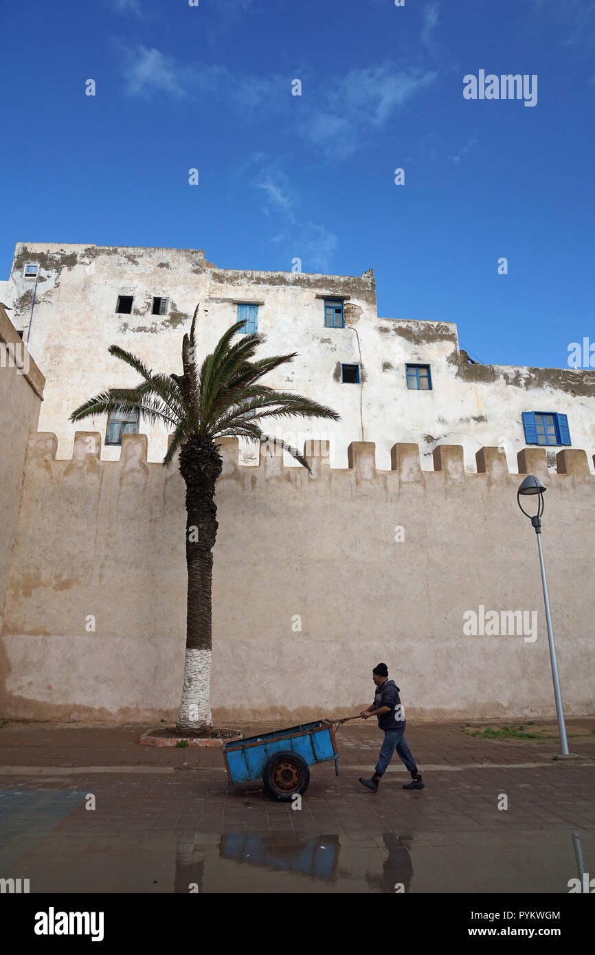 Man with wheel barrow walking along the city wall of Essaouira, Morocco Stock Photo