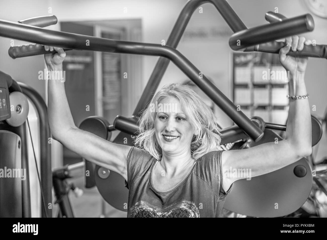 Portrait of smiling female athlete working on seated shoulder press machine. Training arms triceps biceps muscles in modern gym. Healthy lifestyle concept. Shot in black and white. Stock Photo
