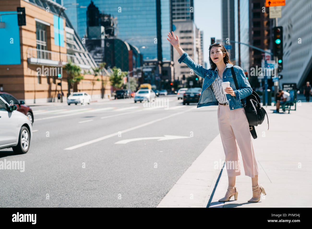 full length photo of beautiful woman beckoning the bus in the busy city. young office lady calling taxi cab in Los angeles, holding coffee standing in Stock Photo