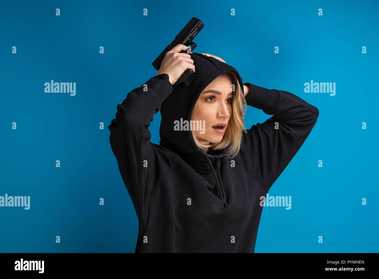 Portrait of hooded girl looking shocked while holding her hands with gun up on the head. Shot against blue background Stock Photo