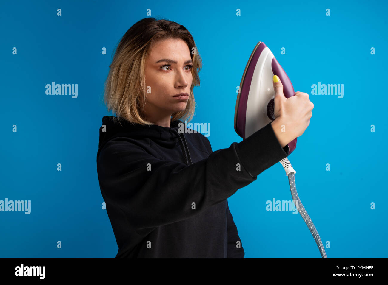 Dark blonde girl making grimaces at a flatiron while holding it raised. Cute commercial idea Stock Photo