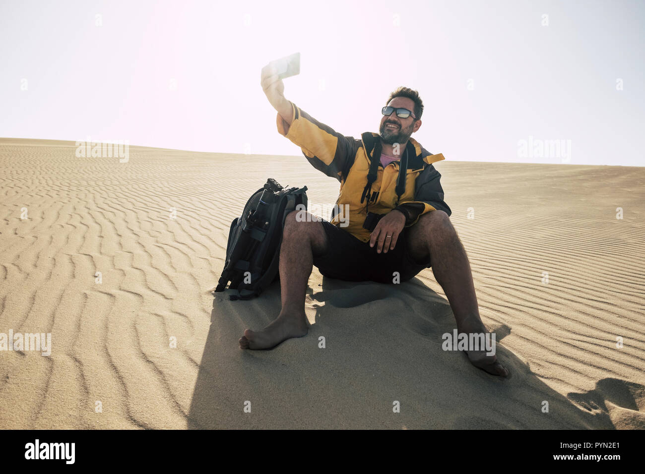Hiker man with camera and backpack is resting sitting on the white sand in the desert by snapping up souvenir photo with smartphon and smiling. concep Stock Photo