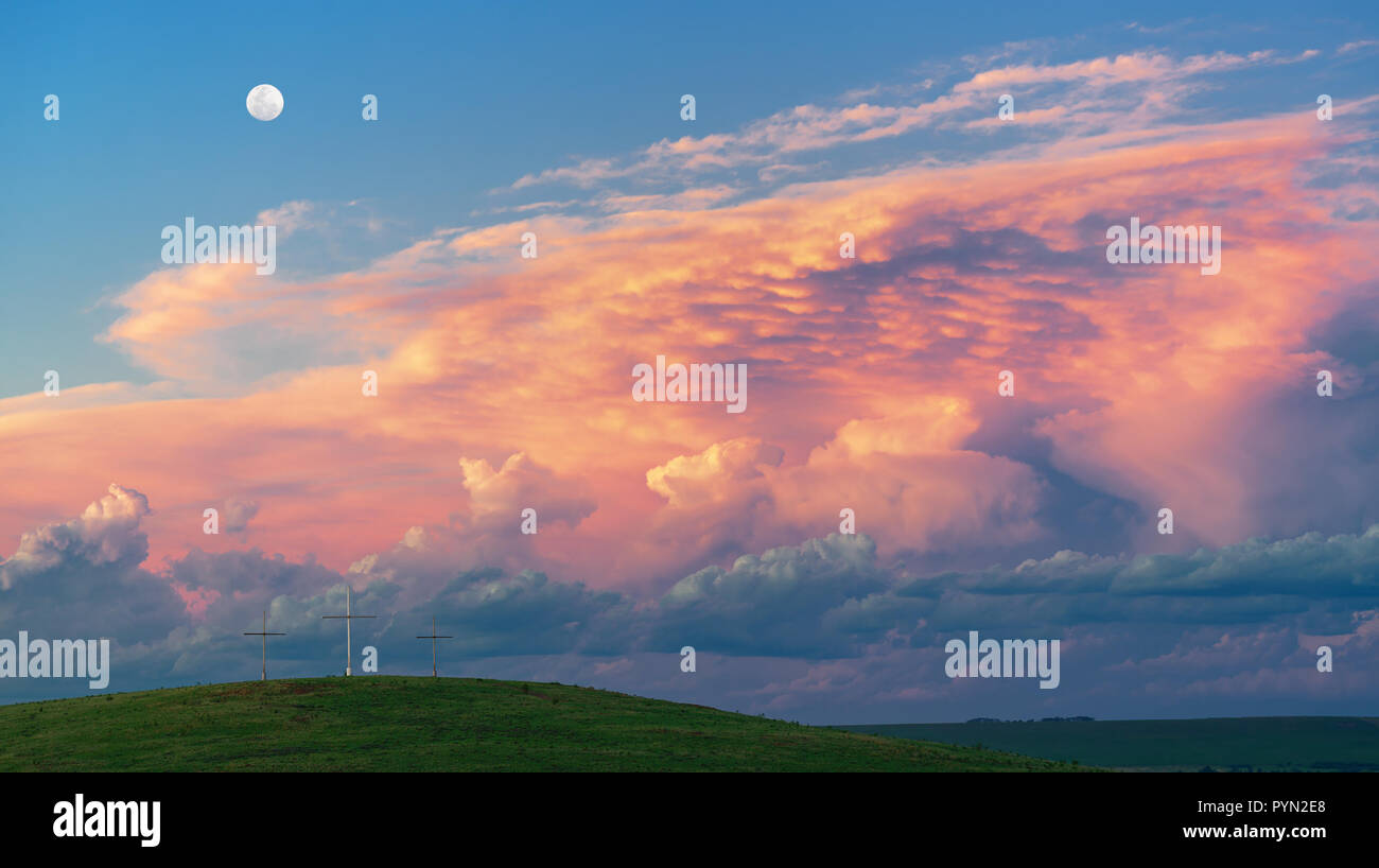 Dramatic clouds filling the sky at sunset. Foreground green hill with three crosses Stock Photo