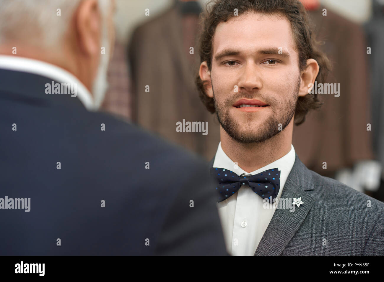 Grey haired man standing back to camera in front of customer. Hadsome young guy in grey jacket and black bow tie in pattern looking at camera. Portrait of attractive brunette with curly hair. Stock Photo