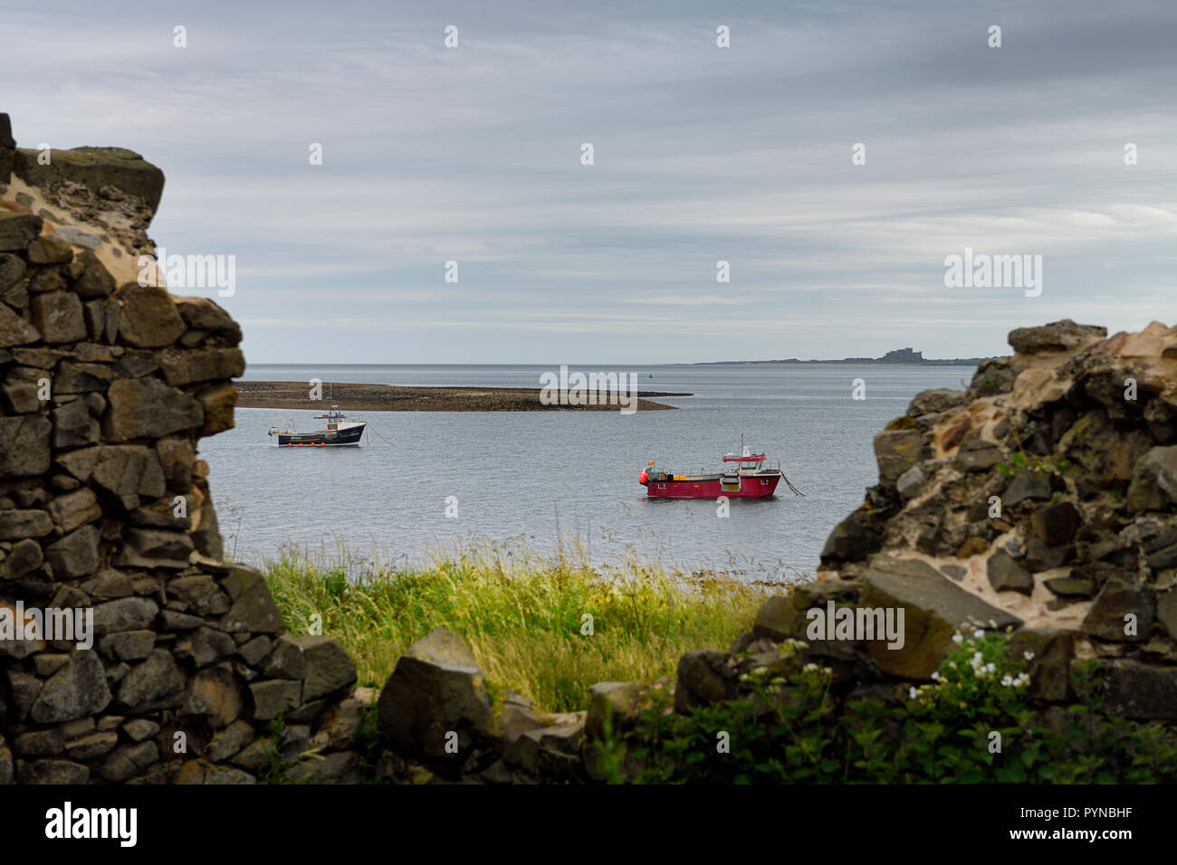 Two fishing boats at low tide in the Harbour of Holy Island of Lindisfarne with Bamburgh Castle through the Steel End ruins England UK Stock Photo