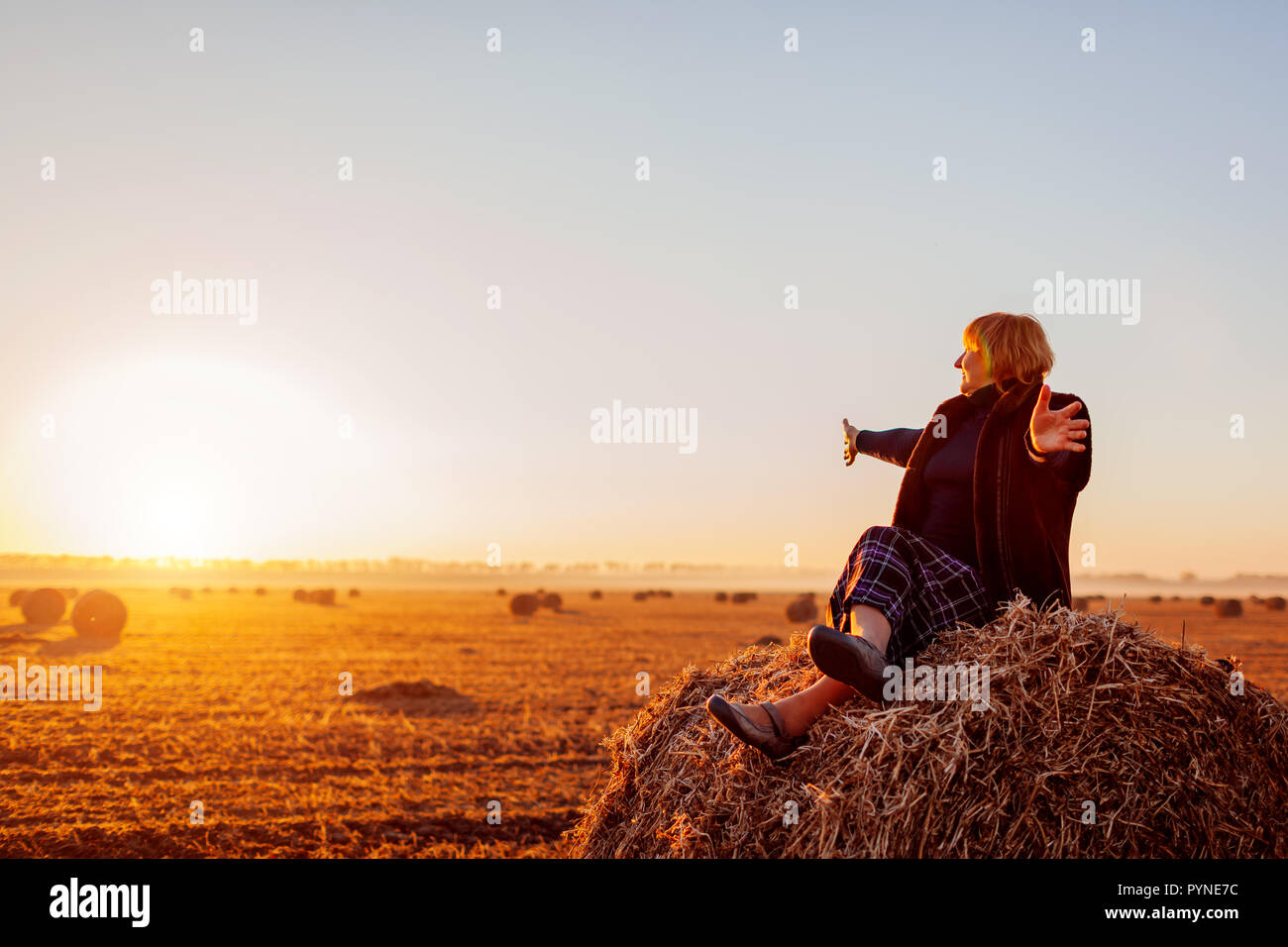 Happy middle-aged woman sitting on haystack in autumn field with arms opened. Relaxing and admiring nature at sunset Stock Photo