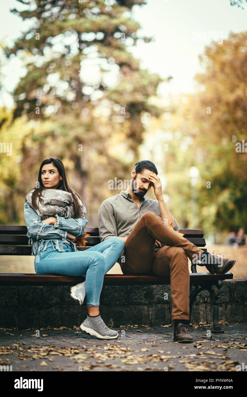 Young couple is sitting in the autumn city park on the bench, quarreling. They are angry and unhappy and not looking at each other. Stock Photo