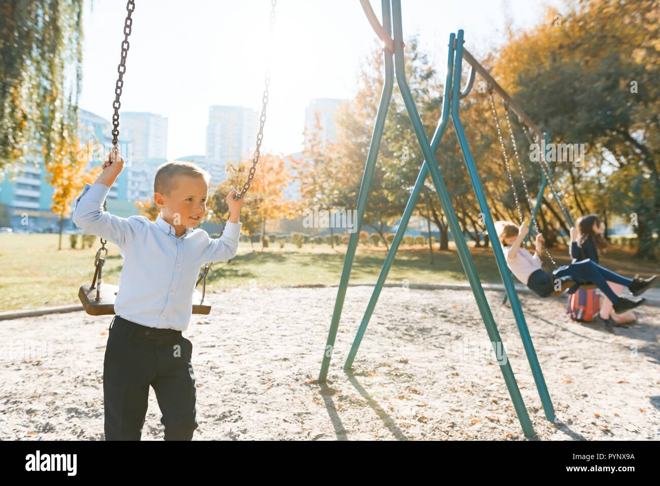 Children ride on a swing in autumn park. Focus on the boy, the girls in the distance, golden hour. Stock Photo