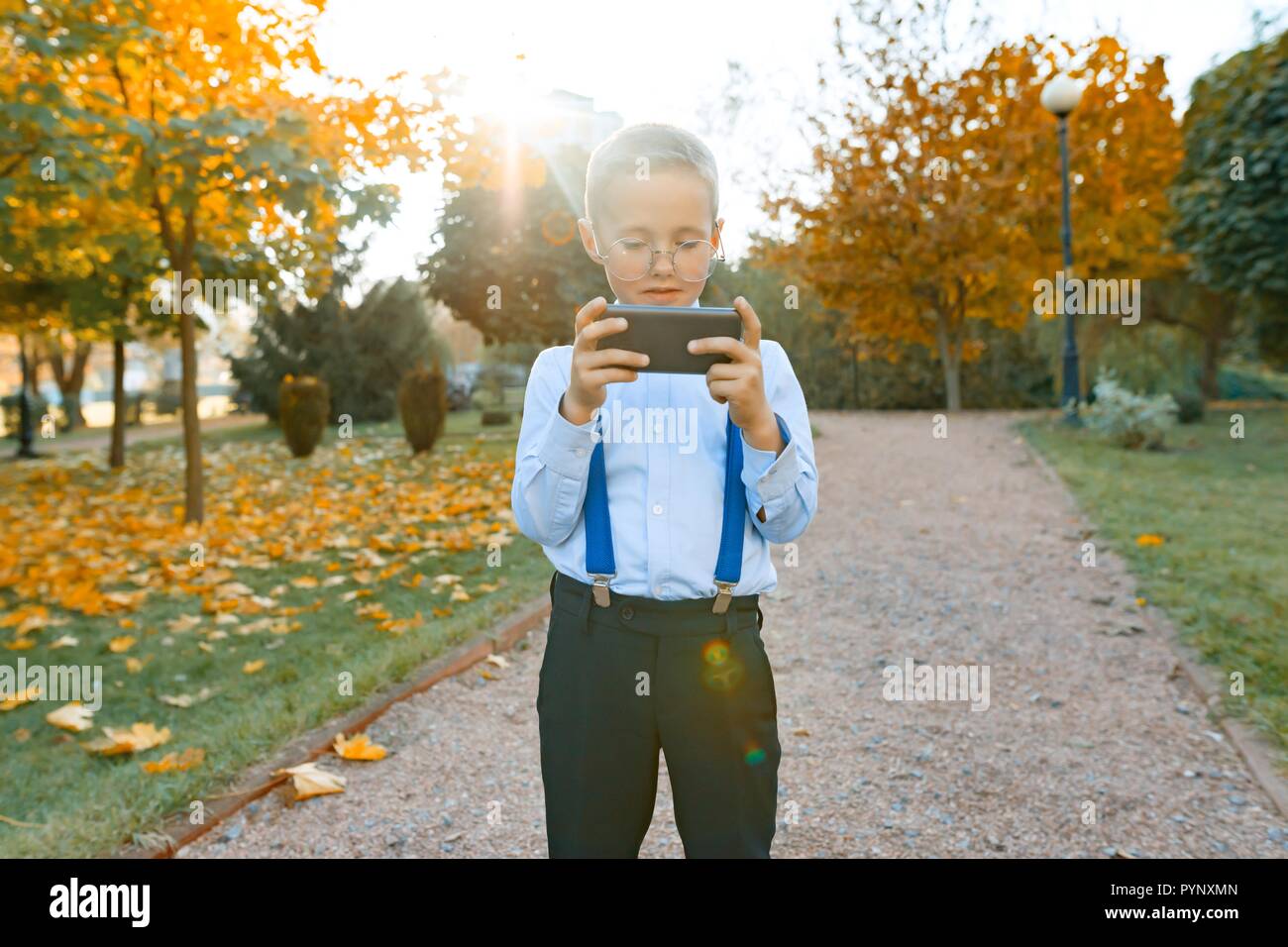 Smart boy in glasses with a mobile phone. A child in a classic shirt in suspenders for trousers, sunny park background, the golden hour. Stock Photo