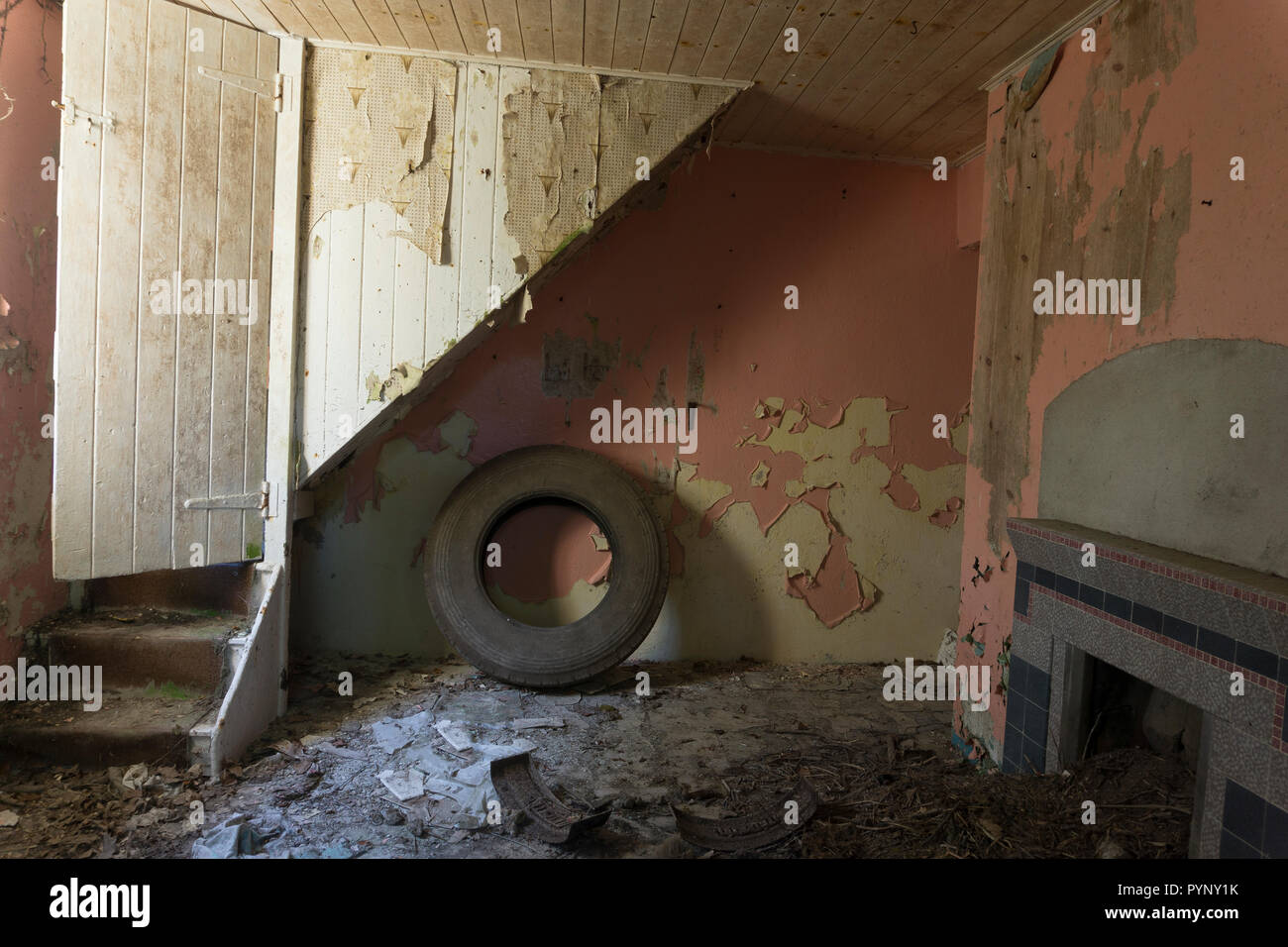 Staircase, with old truck tyre underneath, in an old derelict cottage. Floor strewn with peeled paint and wallpaper, with old fireplace to one side. Stock Photo