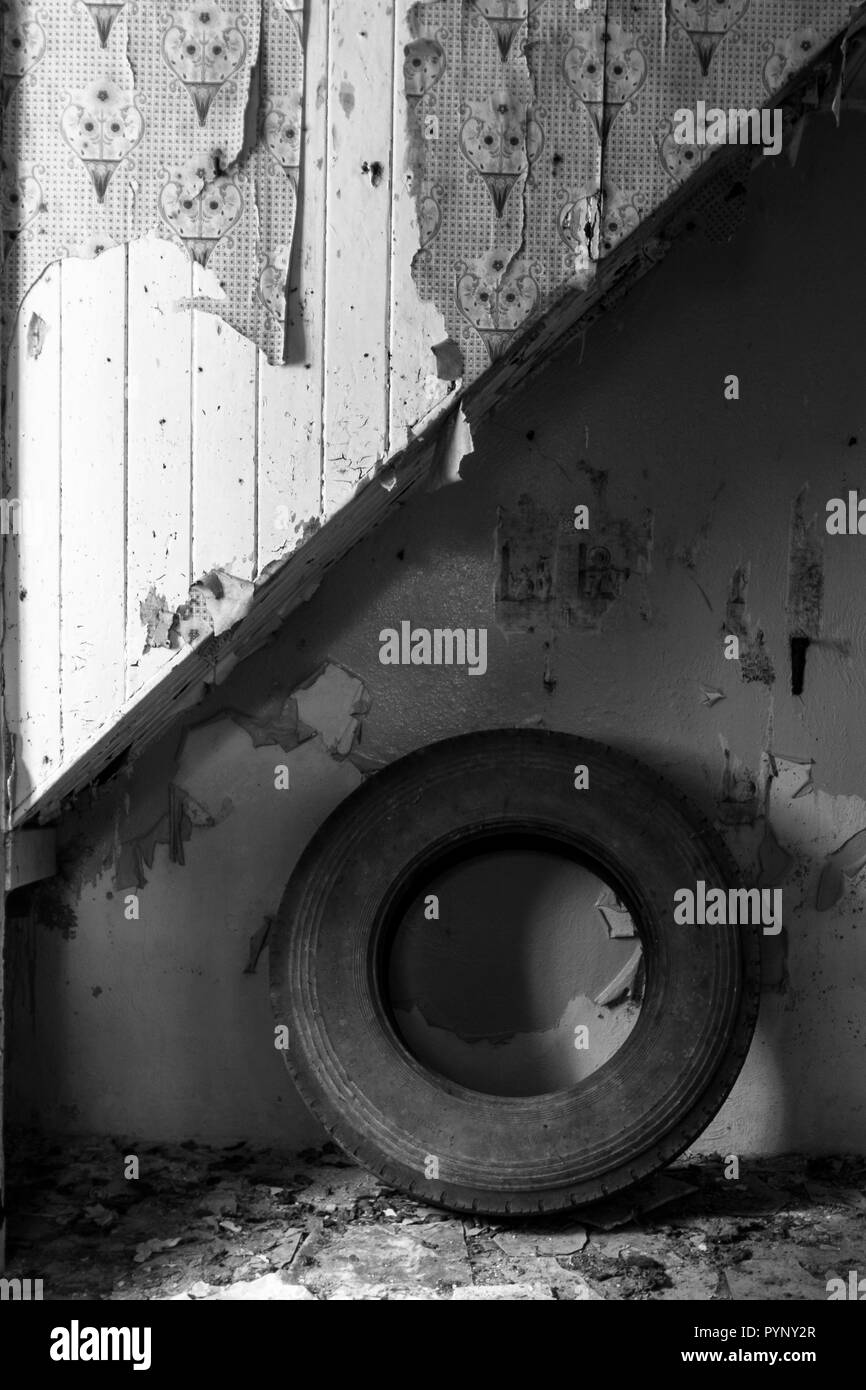 Staircase in old derelict cottage with old truck tyre underneath it.. The floor is strewn with fallen paint and old wallpaper. Stock Photo