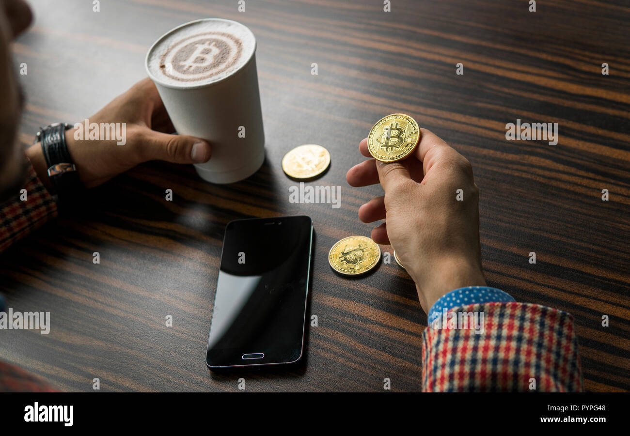 Rich man holding a bitcoin coin and drinking a coffee with a phone and gold  coins of bitcoin on a table. Profit from mining of bitcoins Stock Photo -  Alamy