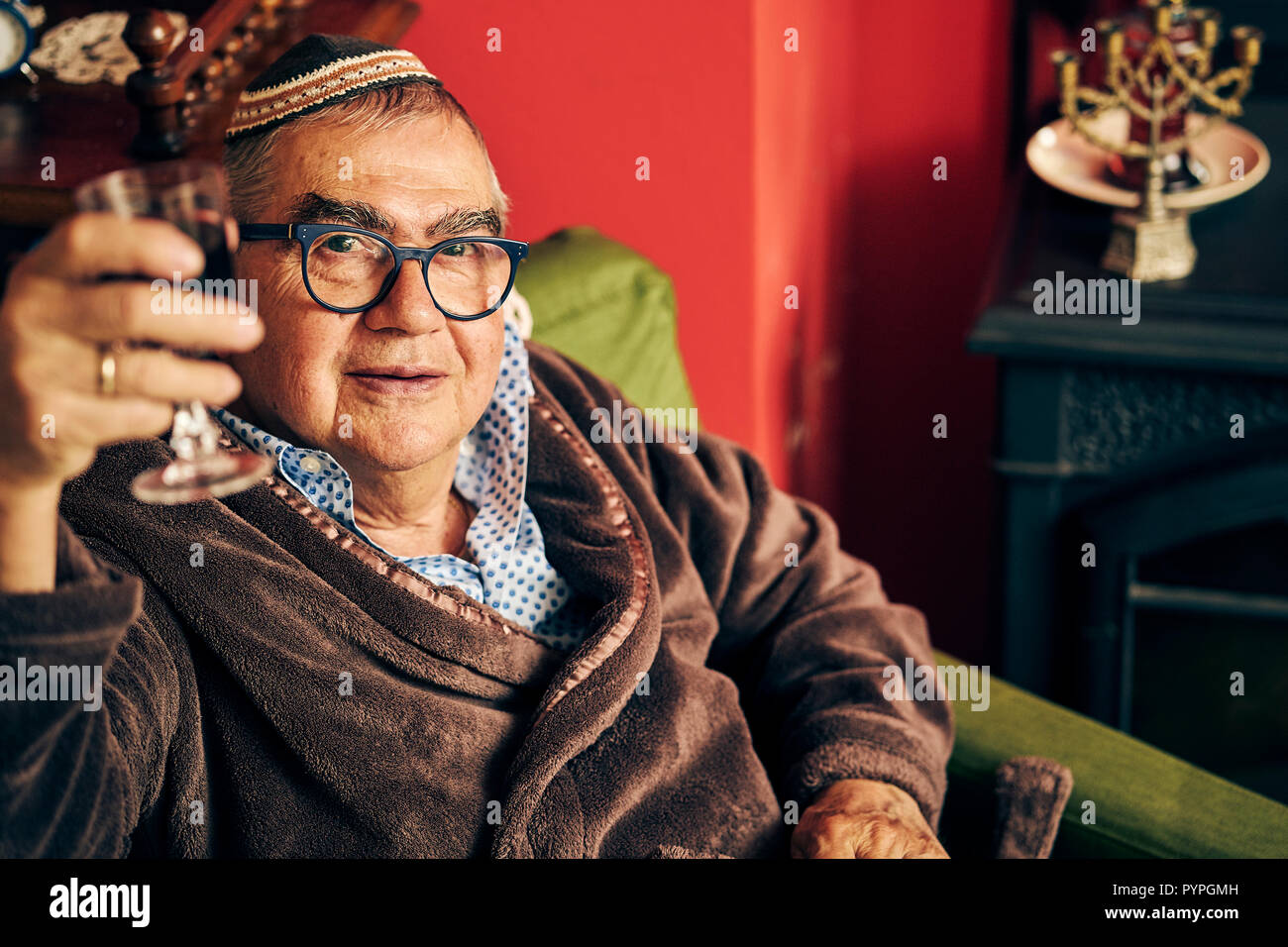 Jewish senior with glasses sitting in the armchair drinking kosher wine ...