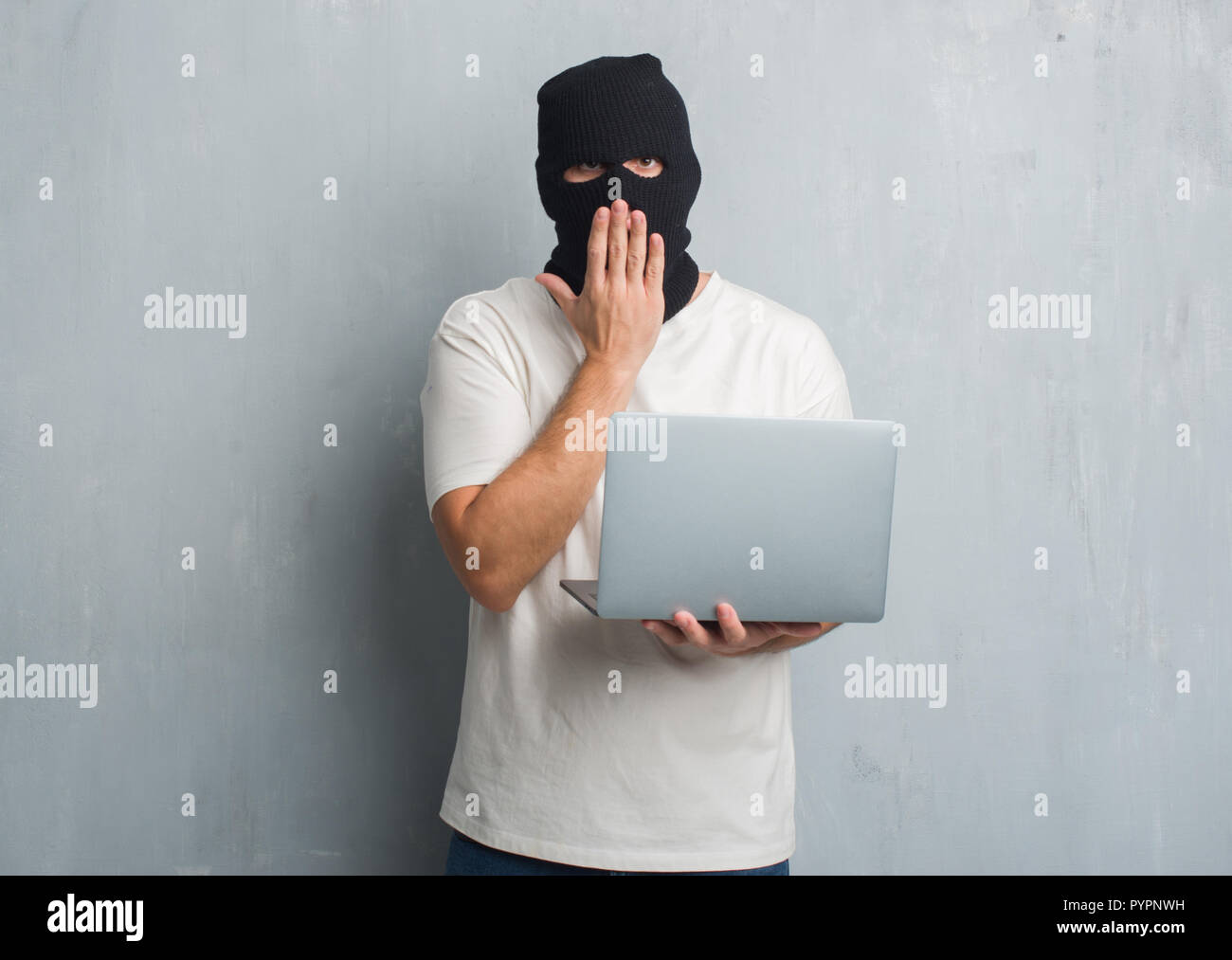 Young caucasian hacker man over grey grunge wall doing cyber attack using laptop cover mouth with hand shocked with shame for mistake, expression of f Stock Photo