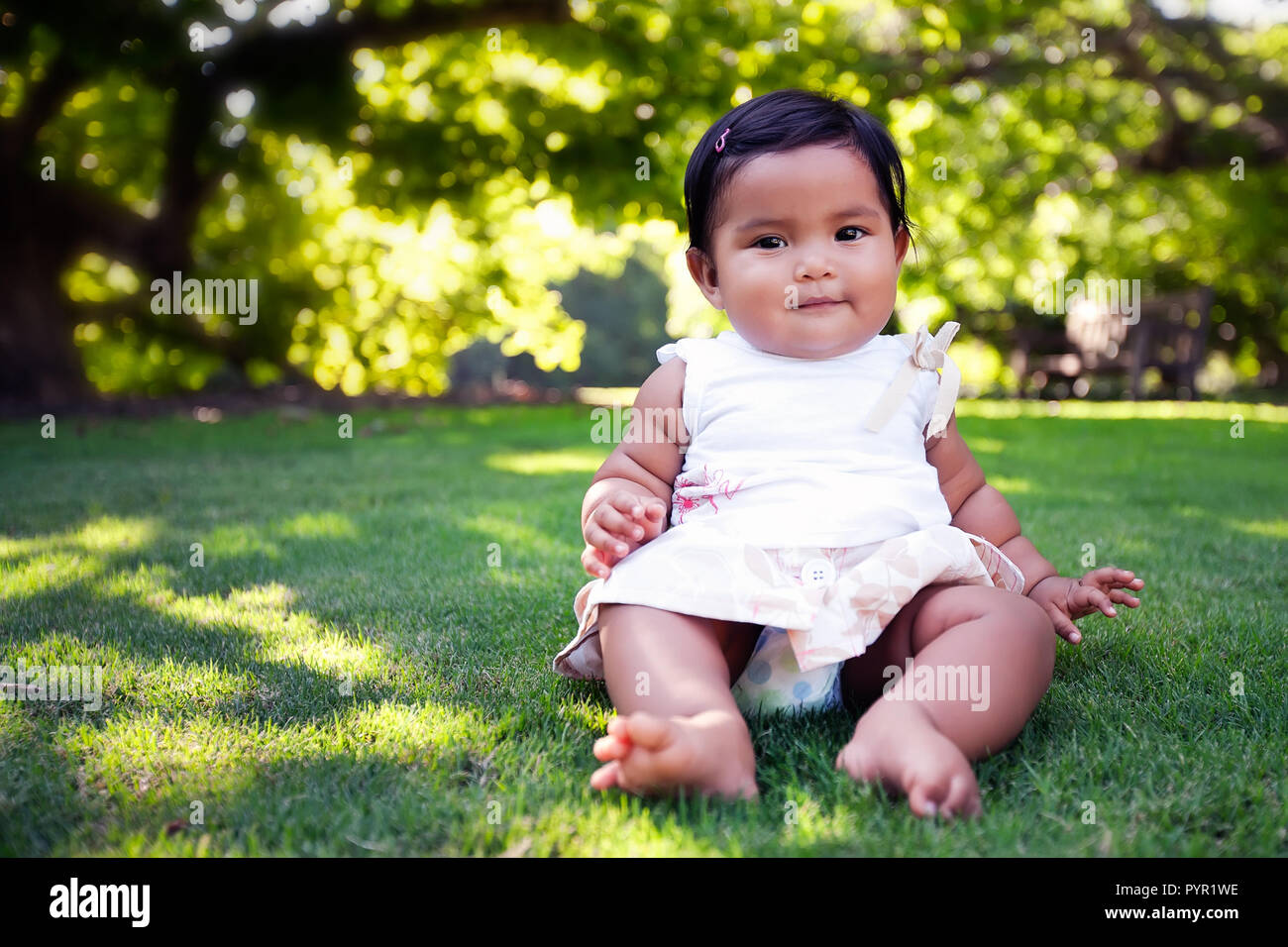 Baby mexican girl with cute smile, an unsupported sitter who has reached a developmental milestone, on a green lawn with beautiful trees. Stock Photo