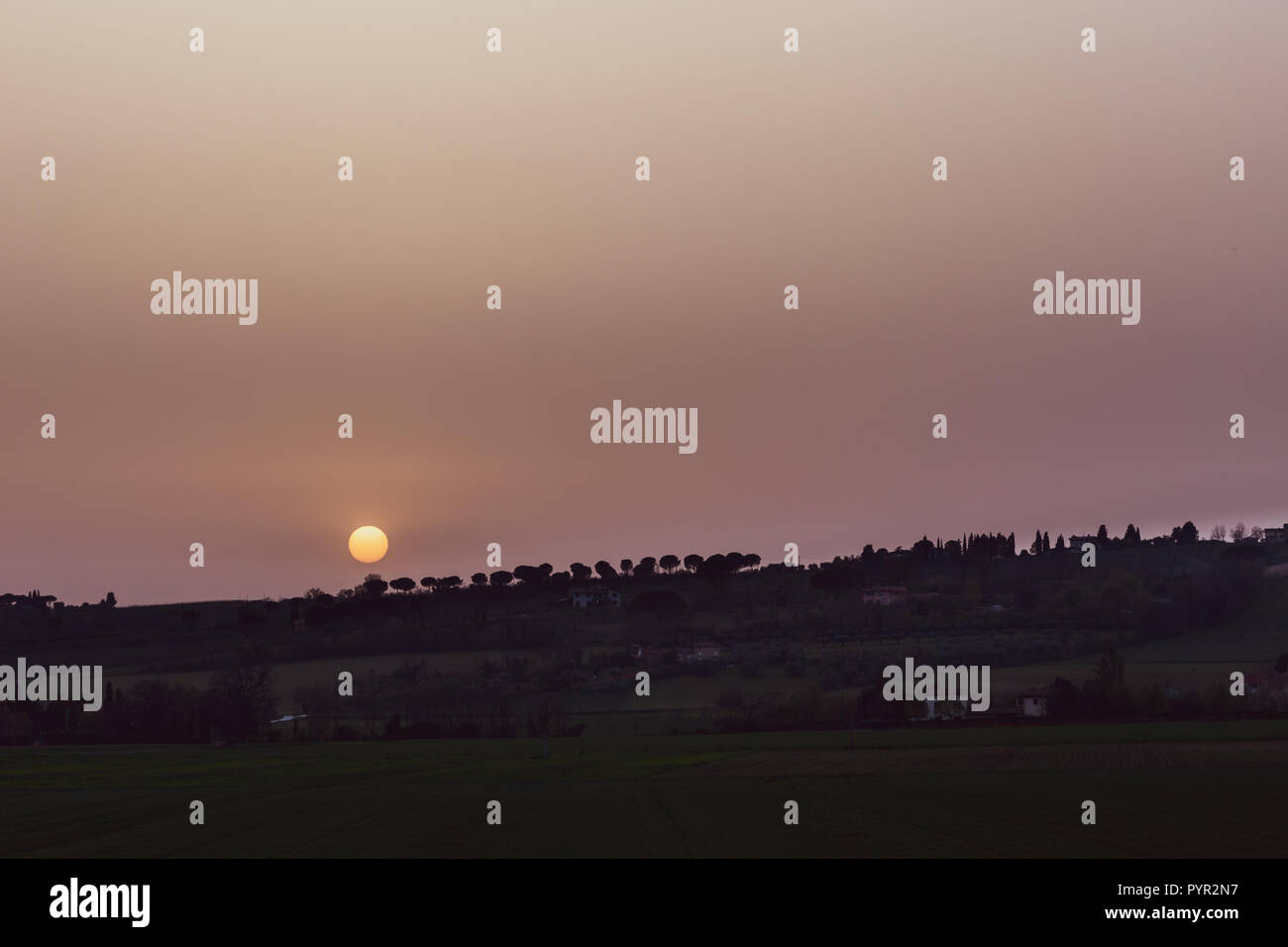 Sunset with sand suspended in the atmosphere, coluring the sky red, over some fields and hills Stock Photo