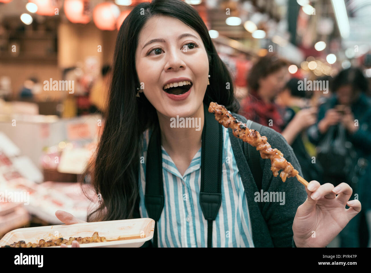 young lady tourist hobby eating local specialty street food in japan. joyful traveler trying barbecue meat in traditional market in osaka in jp. woman Stock Photo