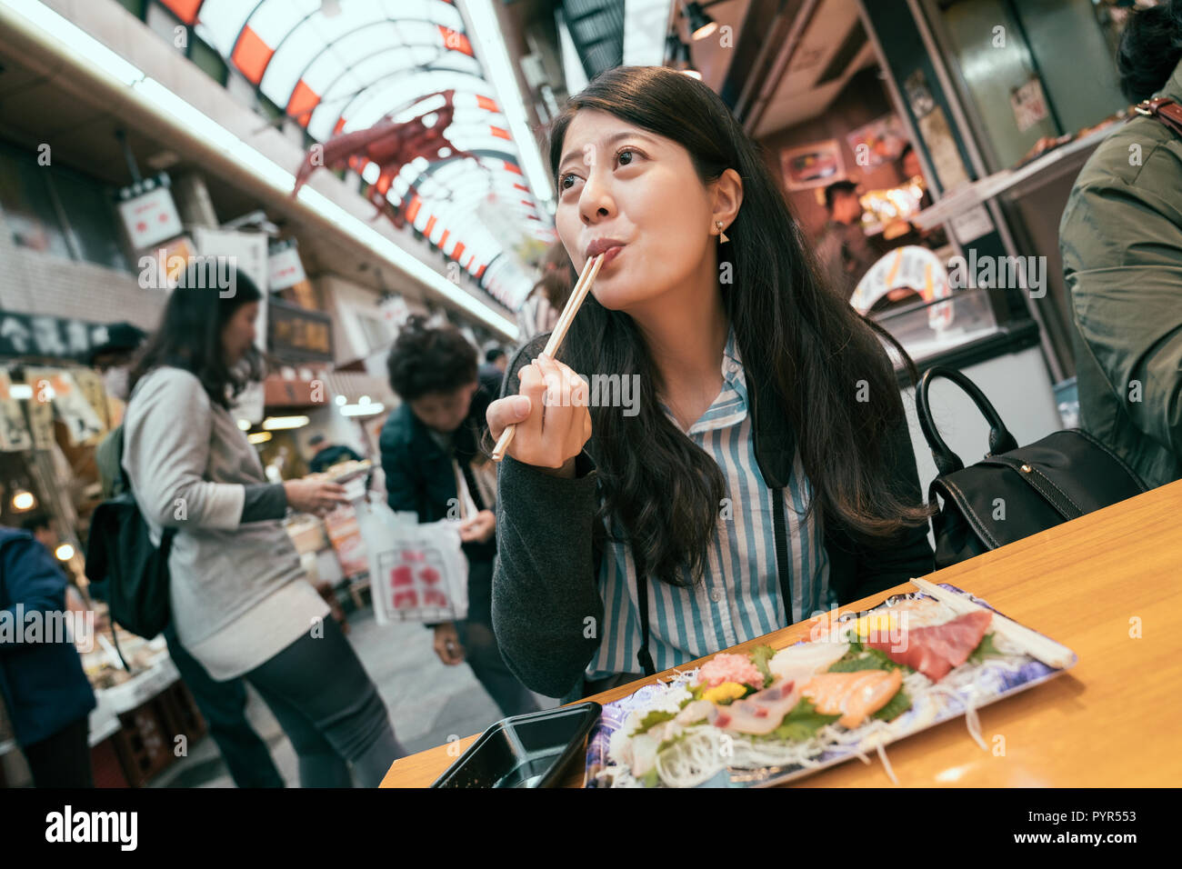 beautiful tourist enjoying sashimi by chopsticks. traveling girl excited eating fresh raw fish in vendor. young traveler sitting in the vendor in indo Stock Photo