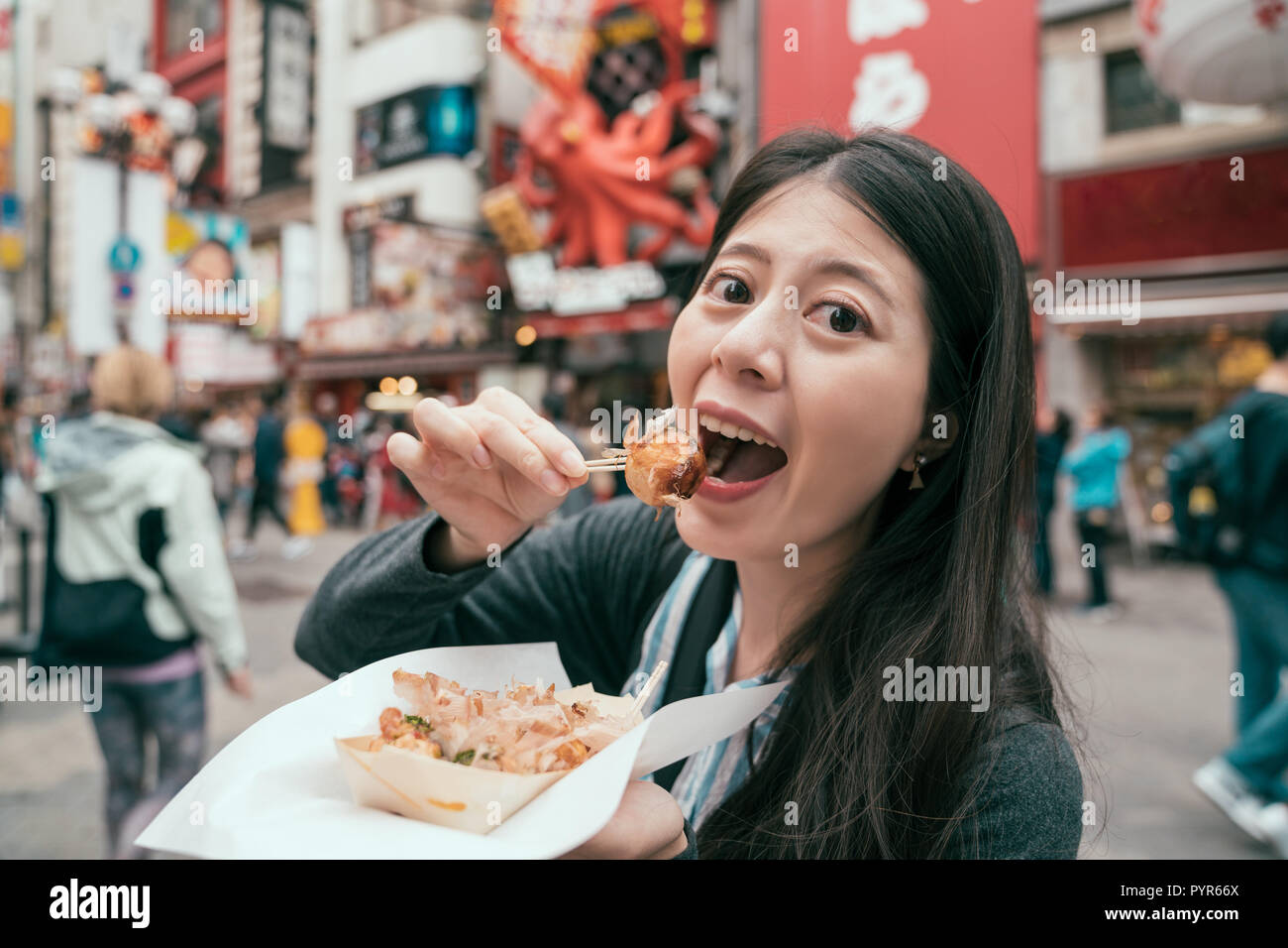 young lady traveler eating takoyaki on the teeming street. girl tourist trying tasty japanese octopus balls on the famous street dotonbori. self-guide Stock Photo
