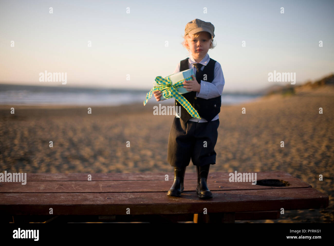 Little boy in old-fashioned clothing carrying books on the beach standing on beach picnic table holding gift Stock Photo
