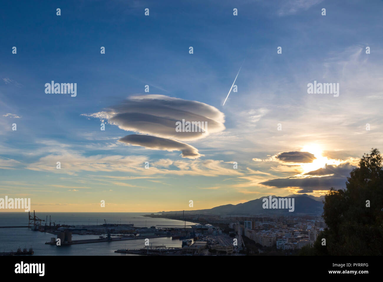 Large circular cloud forms (Altocumulus lenticularis duplicatus V-39) in the evening sky over Malaga city, Spain. Airplane trace in the sky on the bac Stock Photo