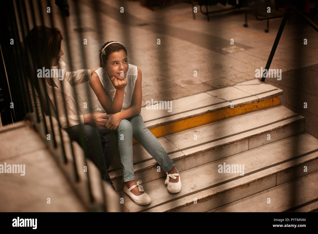 Young girl sitting on the stairs inside a mall with her mother. Stock Photo