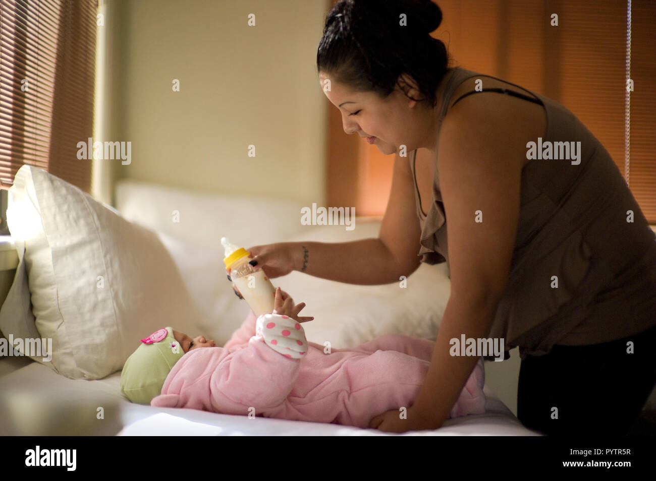 Young mother handing a bottle of milk to her baby daughter who is lying down on the bed. Stock Photo