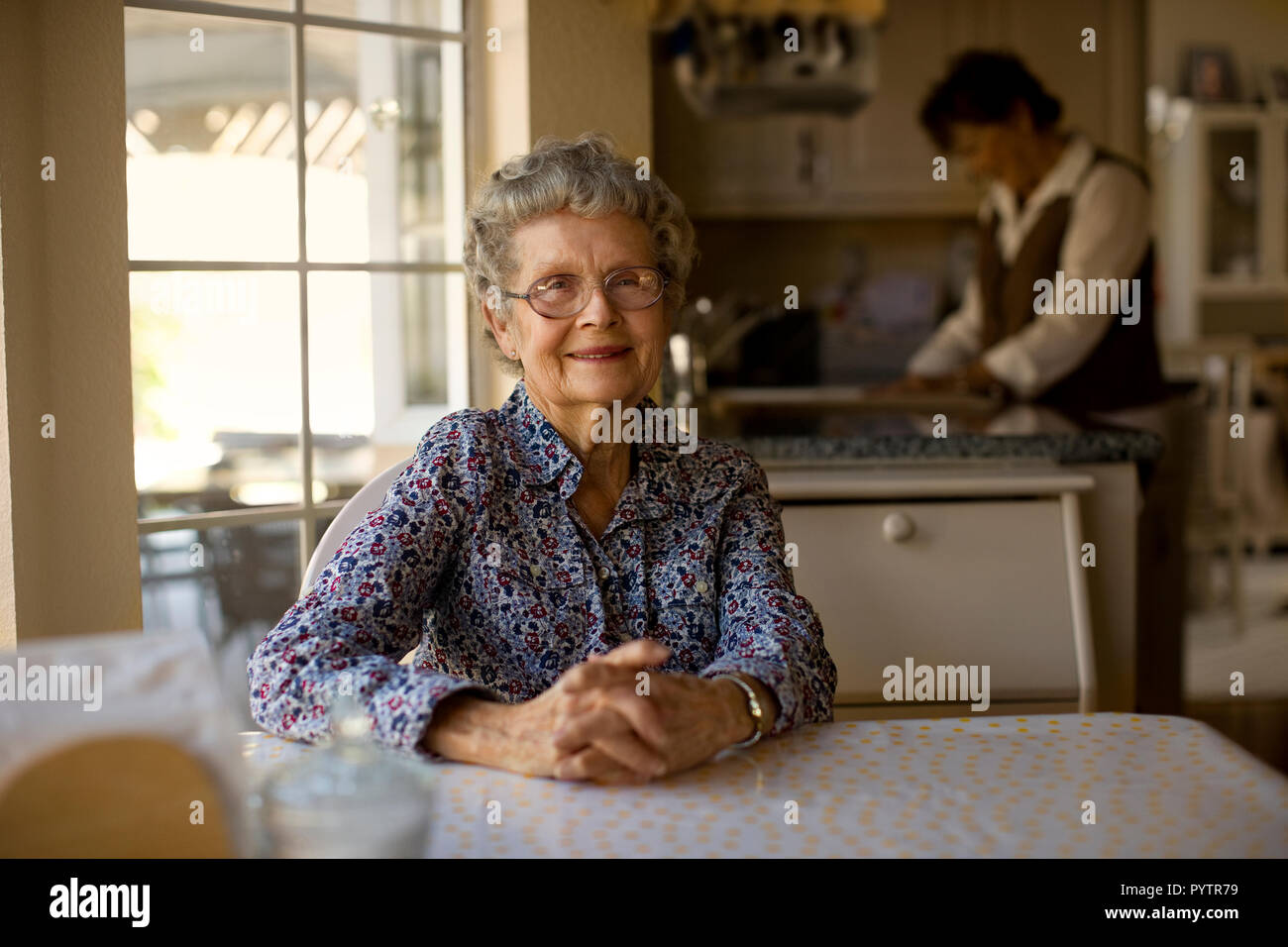 Portrait of a cheerful elderly woman sitting at her kitchen table while her daughter helps out with the dishes. Stock Photo