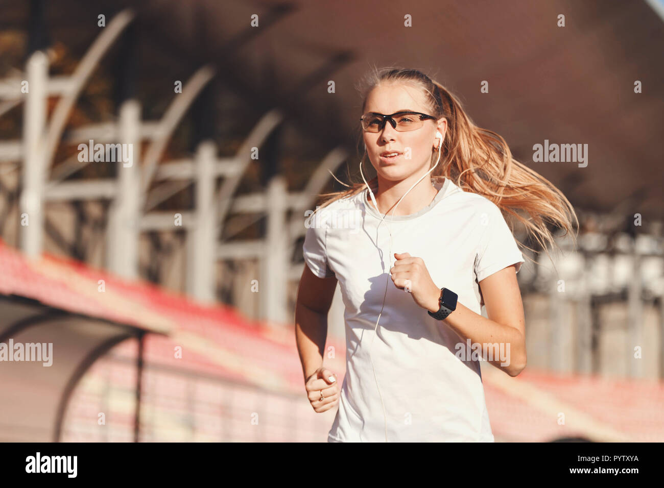 Fitness blonde long haired woman in sportswear listening music and running at the football stadium outdoor, healthy lifestyle and people concept Stock Photo