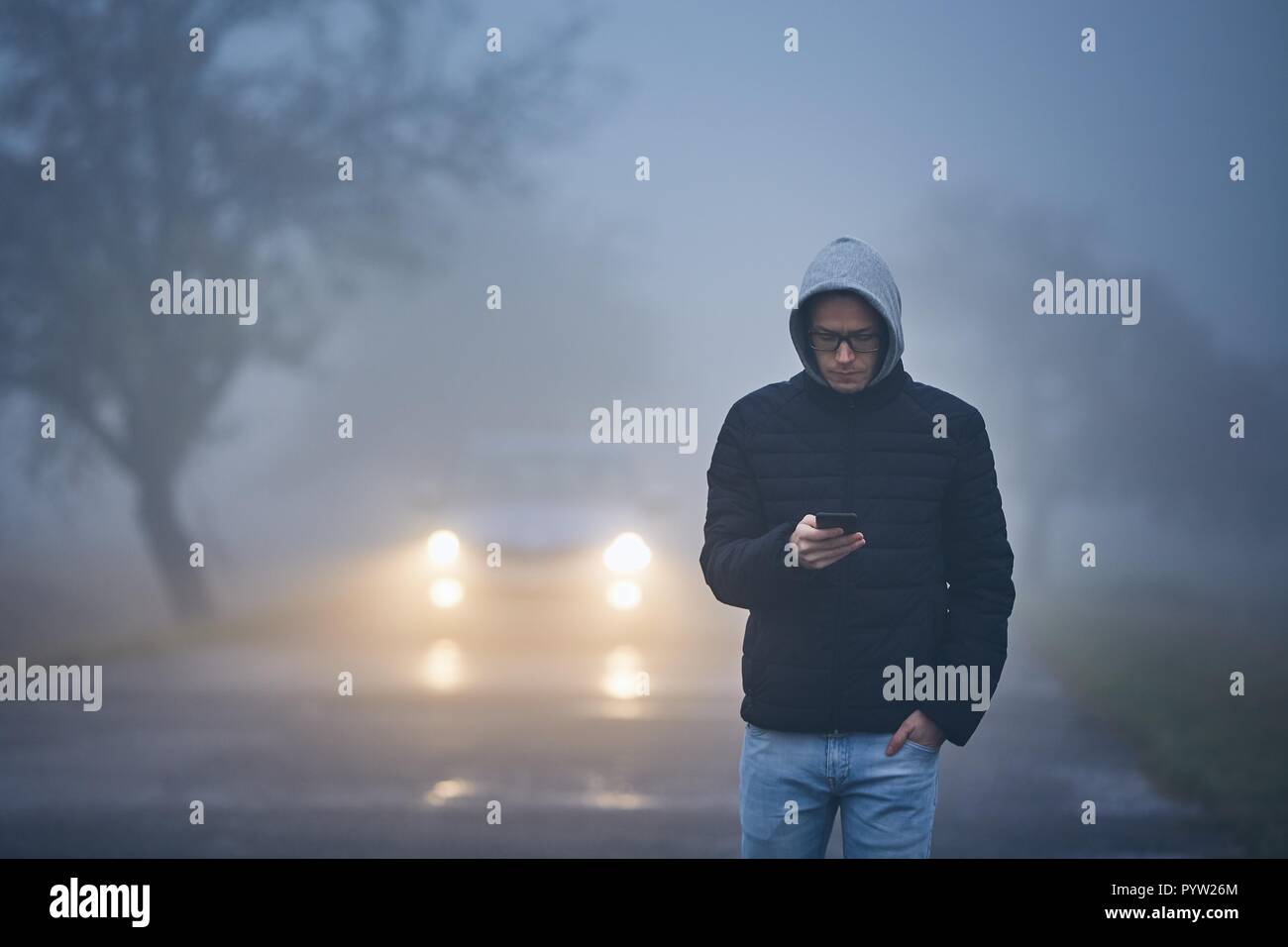 Young man walking on road and using phone. Car approaching in thick fog. Stock Photo