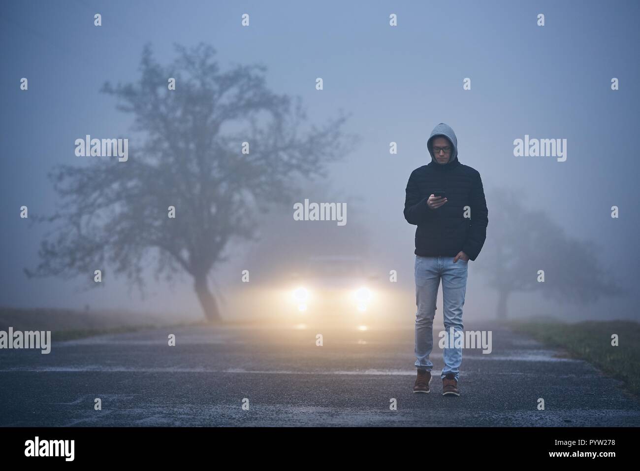 Young man walking on road and using phone. Car approaching in thick fog. Stock Photo