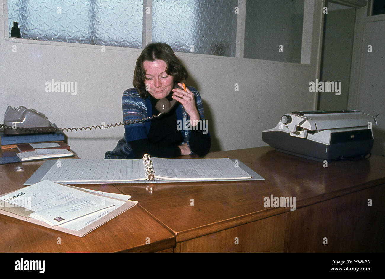 1970s, historical, a female office worker, perhaps an accounts clerk, sitting at her desk looking at numbers in a clip file speaking on the telephone, England, UK. Stock Photo