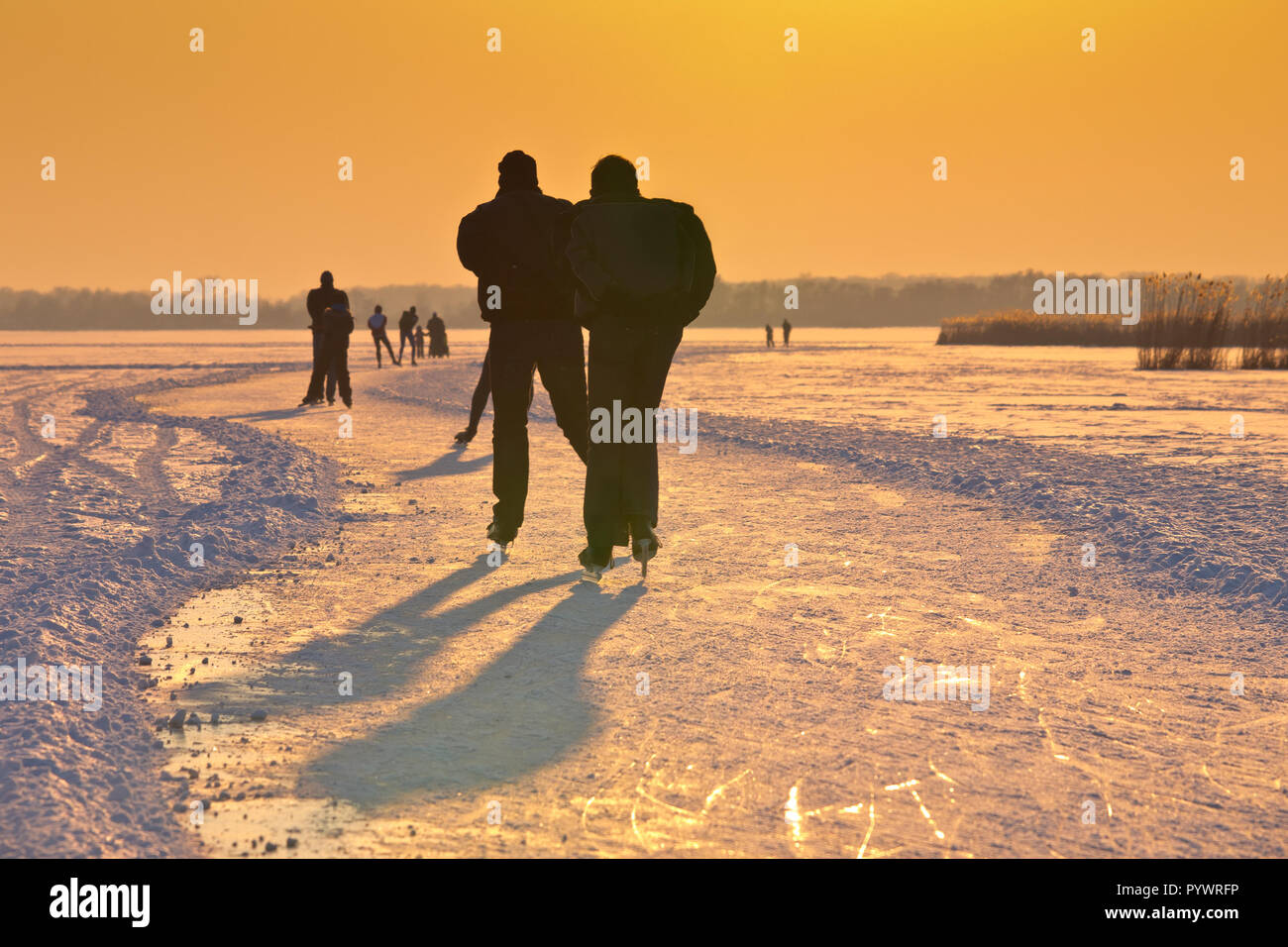 Ice Skaters on frozen lake seen on their back under orange setting sun Stock Photo