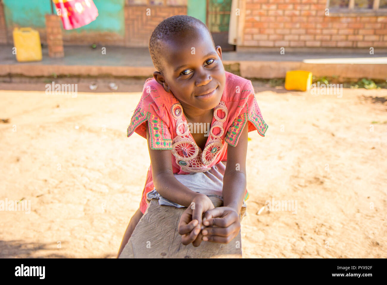 A beautiful young Ugandan black girl leaning on a wooden bench. She has a serene yet happy look. She is dressed a pink ornamented T-shirt. Stock Photo