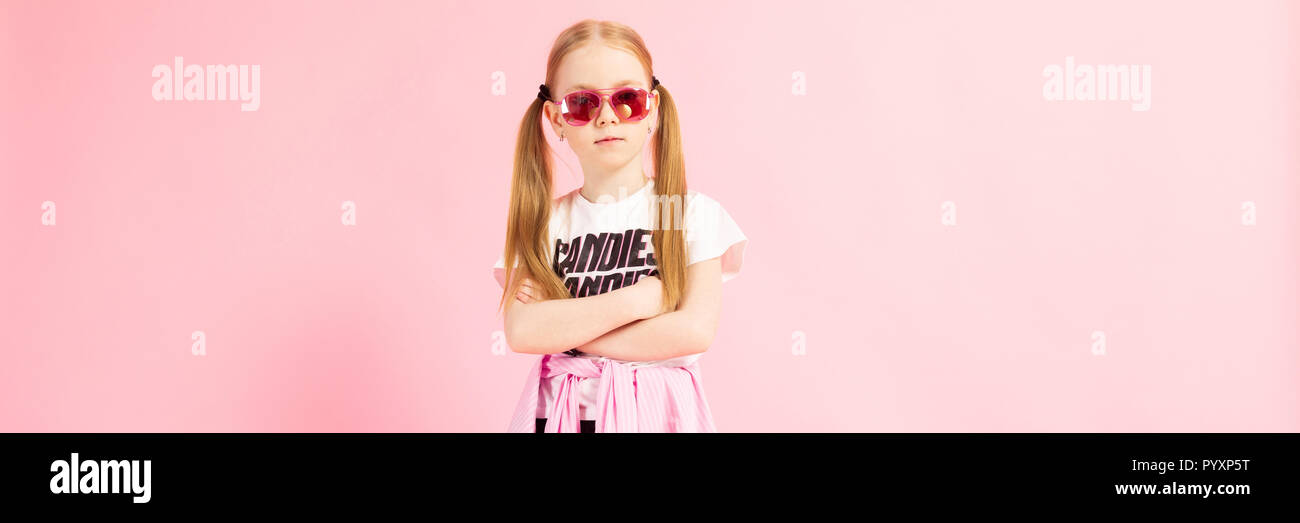 Girl with red tails on a pink background. A charming girl in bright sports clothes and pink glasses folded her arms on her chest. Stock Photo