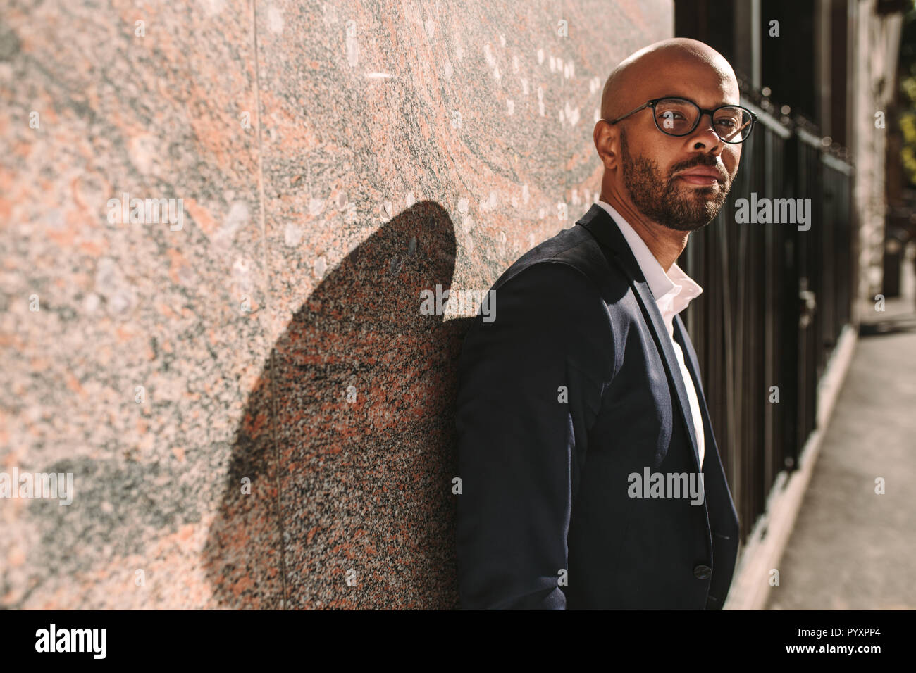 Portrait of handsome young man wearing suit leaning to a wall outdoors and looking at camera. African businessman with beard wearing eyeglasses relaxi Stock Photo