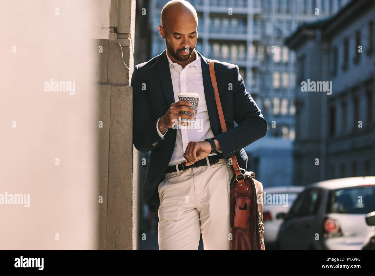 Young african businessman leaning to a wall outdoors and checking time on his wristwatch. Man in suit waiting for someone outdoors in the city. Stock Photo