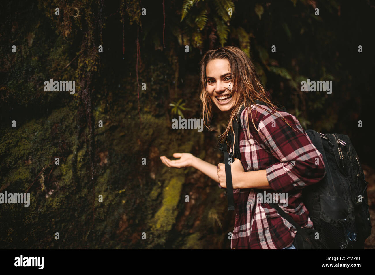 Portrait of beautiful young woman putting her hand under the water coming over from the rocks in forest. Cheerful woman enjoying a hike in rain forest Stock Photo