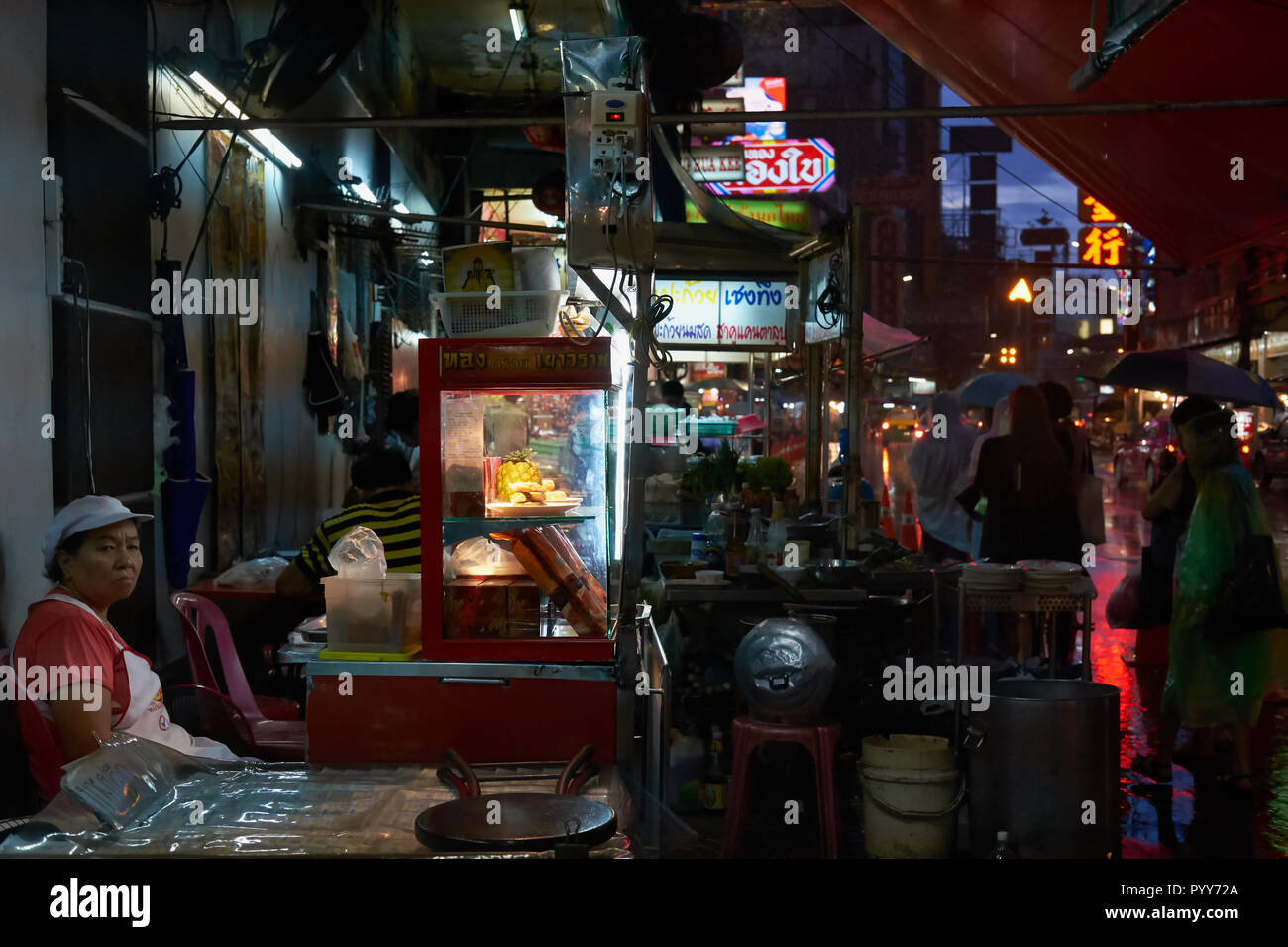 On a rainy monsoon evening in Chinatown, Bangkok, Thailand, a food vendor sits at her clustered stall and gloomily looks out for customers Stock Photo