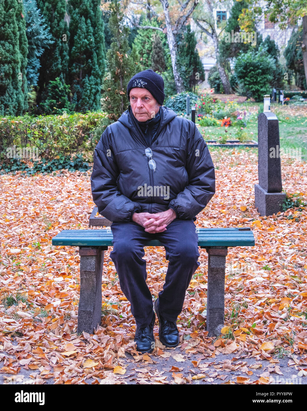 Senior man sitting on decorative wrought iron & wood bench, in cemetery -  looking pensive,thoughtful, sad. Stock Photo