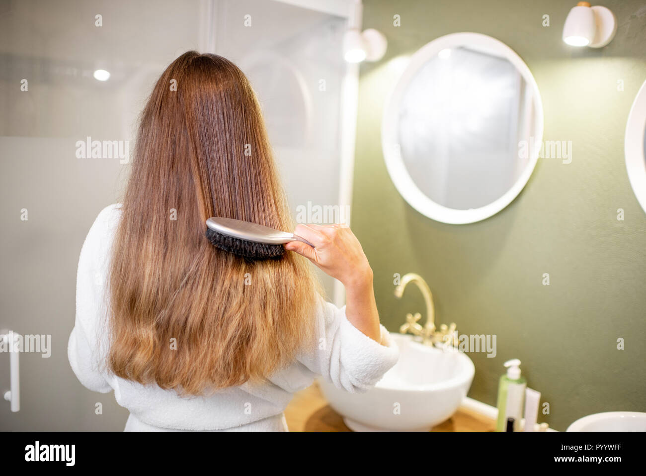 Woman in bathrobe combing hair with brush in the bathroom, rear view Stock Photo