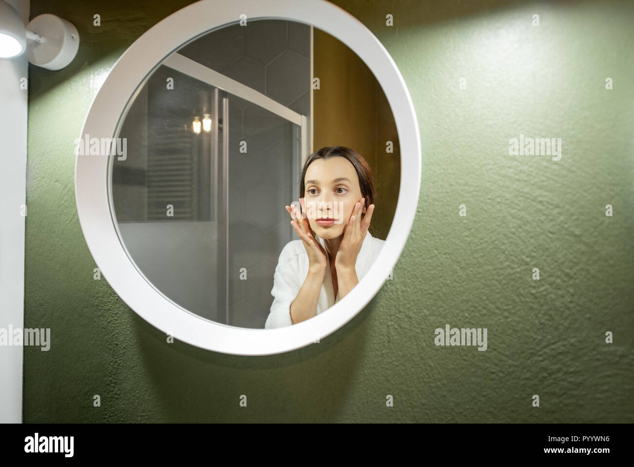 Woman In Bathrobe Making Facial Massage Looking Into The Round Mirror In The Bathroom Stock