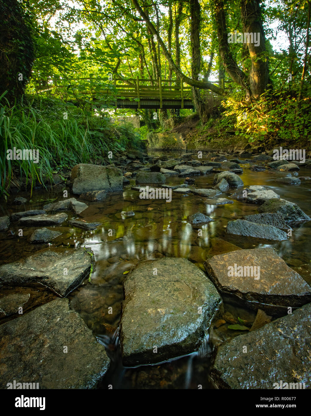 rocks in a very calm stream with a wooden bridge passing over Stock Photo