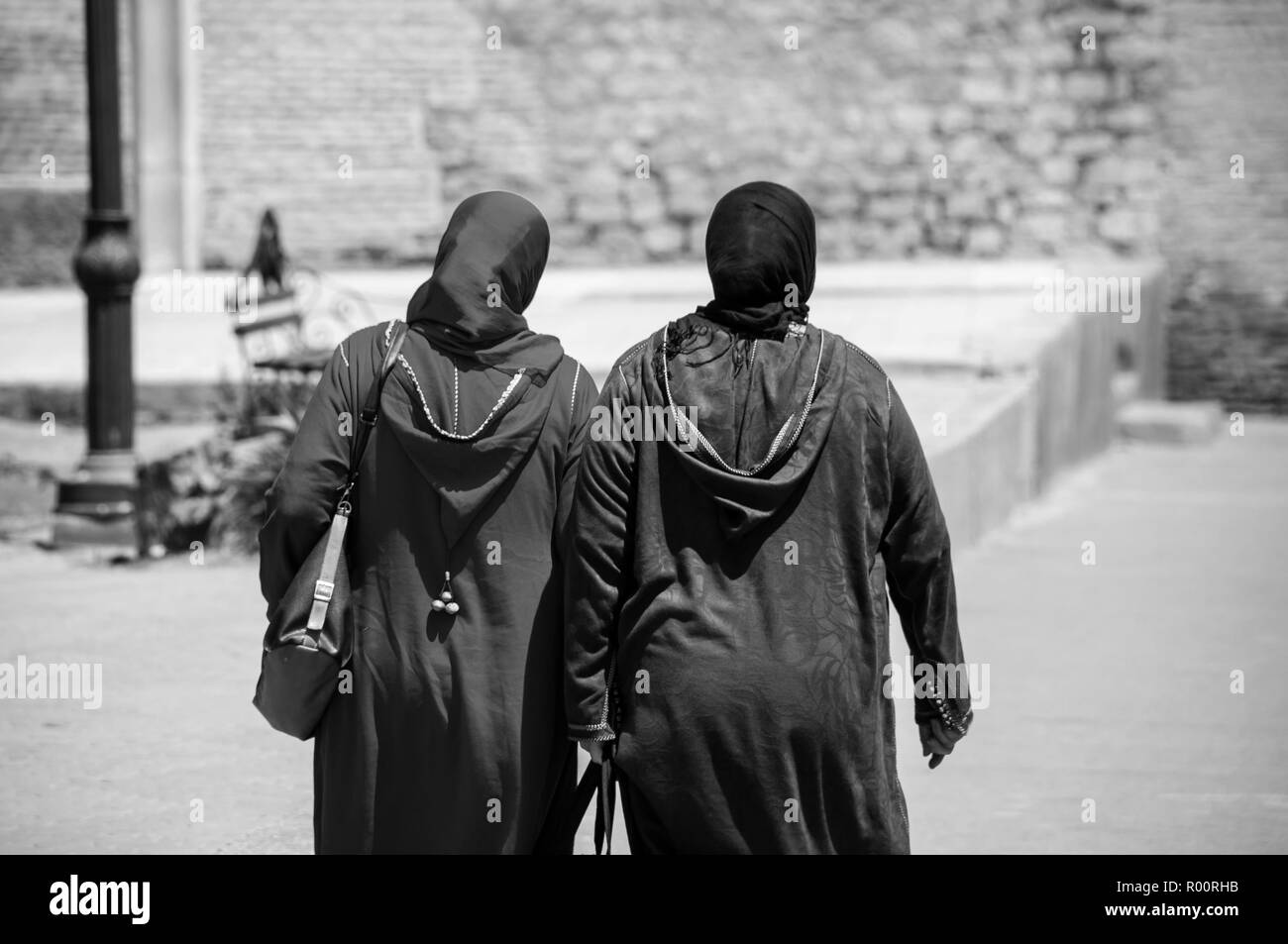 Two moroccan women seen from the back dressed in typical blue and brown djellaba walk down the streets of Marrakesh Medina, Morocco. Black and white Stock Photo